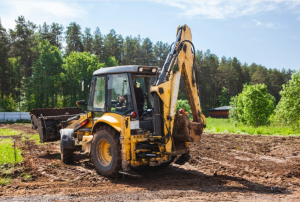 Yellow land-clearing machine removes vegetation and debris for defensible space work on a dirt site, surrounded by green trees and blue sky.